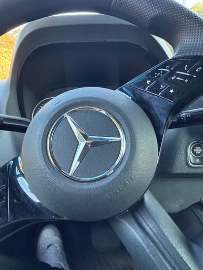 The image depicts the interior of a modern Mercedes-Benz vehicle, located in the city of Celle, Germany. The perspective is from the driver's seat, focused on the steering wheel and a portion of the dashboard.
The central object is a black, multi-function steering wheel with a prominent chrome Mercedes-Benz three-pointed star logo encircled on its airbag cover, which also bears the embossed word "AIRBAG." The right spoke of the steering wheel features a glossy black panel with multiple illuminated or marked control buttons for functions such as navigation, volume, phone, and a star/favorite button. Behind the steering wheel, a dark, unlit digital instrument cluster is visible within the dashboard housing. To the right of the steering wheel, on the dashboard, a circular "START ENGINE STOP" button is present, accompanied by an "A" symbol, likely indicating an automatic start/stop system.
In the lower-left corner of the frame, a person's left foot, clad in a white athletic shoe, is visible resting within the vehicle's footwell, suggesting the presence of an occupant in the driver's position. Through the windshield at the top of the frame, blurry yellow/orange foliage and a bright sky are discernable, indicating an outdoor, daytime setting with natural light illuminating the interior. Reflections are visible on the glossy surfaces of the steering wheel controls and the Mercedes logo.