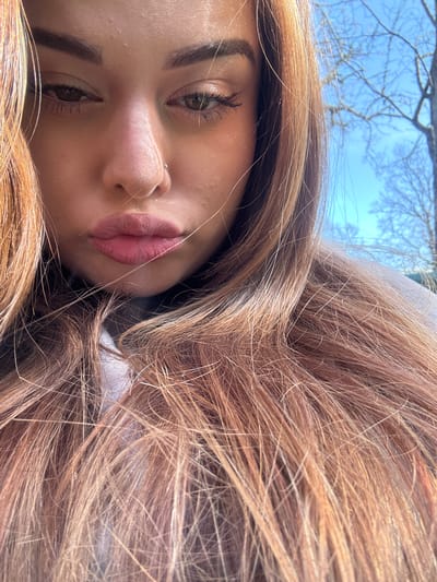 The image presents a close-up, slightly angled portrait of a woman. Her face is the primary subject, featuring light skin, prominent eyebrows, and eyes with visible eyelashes. She has a nose piercing and lips with a distinct color. Her hair, a blend of brown and blonde shades, surrounds her face. The background shows a glimpse of a blue sky and bare tree branches. There is no evidence to suggest that this picture was taken in Gorno Trape, Bulgaria, as the context does not indicate the location. The environment is unidentifiable.