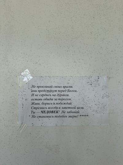 A close-up shot captures a piece of paper, likely a handwritten or printed note, taped to a mottled, off-white wall. The paper is affixed with clear, crinkled tape. The text on the paper is in Russian and appears to be a poem or a set of maxims. The wall itself is textured and speckled with what looks like mold or grime, creating a speckled, uneven appearance. The lighting suggests an indoor or shaded outdoor environment, with no distinct shadows indicating direct sunlight. The image provides no contextual clues about the specific location within Podporozhye, Russia, beyond the presence of the text and the wall.