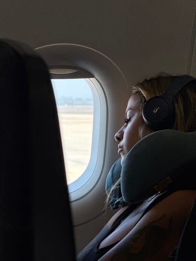 A woman wearing headphones and a neck pillow looks out the airplane window. The window frame is white and the view outside is blurry, but appears to be a bright, light-colored landscape. The woman's face is partially visible, showing her eye, nose, and mouth. A tattoo is visible on her arm. The seat in front of her is dark and out of focus. The overall lighting is soft and diffused.