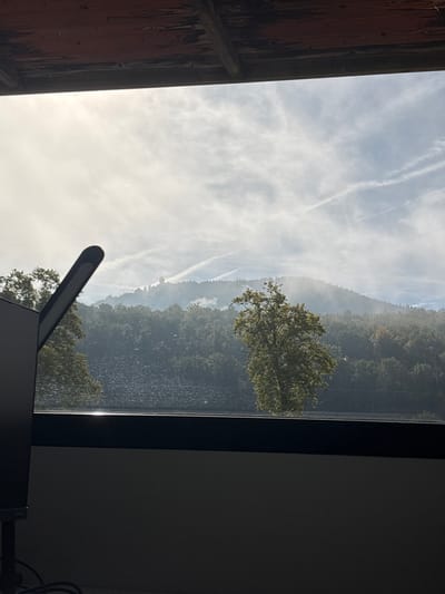 The image displays an outdoor landscape viewed from an interior vantage point through a window in Lièpvre, France. The foreground contains a partial view of a black monitor screen on the lower left, a black desk lamp angled upwards to the right, and a dark horizontal window frame or sill reflecting a strip of light. Below the frame, a dark, monochrome interior wall is visible. Above the window, a reddish-brown wooden ceiling structure with exposed beams is present.

Beyond the window, a dark body of water with visible ripples extends across the midground. Immediately beyond the water, a dense green forest covers a sloping hillside. Patches of white mist or low-lying fog partially obscure sections of the forest, particularly on the upper slopes. On the highest distant hilltop, the Haut-Koenigsbourg Castle, characterized by its distinctive towers, is visible, partially veiled by the surrounding mist. The sky is bright and hazy on the left, featuring scattered cirrus-like clouds and multiple linear aircraft contrails. No individuals or active subjects are depicted.