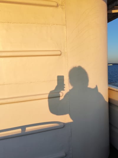 The image shows the shadow of a person holding a smartphone, taking a selfie. The shadow is cast on a light-colored, textured wall, possibly on the exterior of a vessel. The wall has horizontal ridges, and a seam runs vertically down the left side. To the right, a sliver of blue sky and the ocean is visible, with a distant boat on the horizon. The lighting suggests it is either sunrise or sunset. The location is identified as Kakkarovo, Russia.