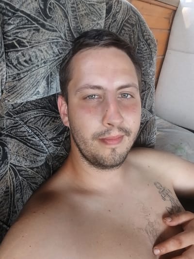 The media file shows a close-up, top-down view of a young man lying on his side on a couch in what appears to be a dimly lit room. The man has light skin, short brown hair, a beard, and light-colored eyes. He is shirtless, revealing a tattoo on his chest. His left hand is visible near his chest. The couch has a dark, patterned fabric. A portion of a light-colored couch or chair is visible in the lower right corner. The lighting suggests it could be daytime but indoors, possibly in the evening. No specific weather or time of day cues are evident. No text is visible in the image. The man's expression is neutral. The setting appears to be a home environment in Vil'nokur'yanivs'ke, Ukraine.