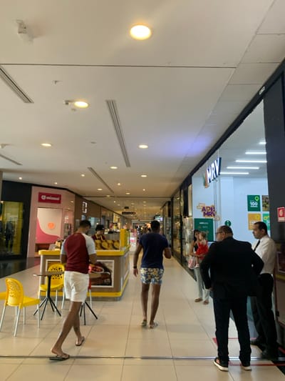 This image depicts an indoor scene within a shopping mall, specifically identified as Shopping Camará in Camaragibe, Brazil. The environment features a wide, brightly lit corridor with a light-colored tiled floor and a high white ceiling equipped with numerous circular recessed lights, linear air conditioning vents, and smoke detectors.
On the left side of the corridor, multiple retail storefronts are visible. Prominently featured is a "miamiau" store with a pink, orange, and white graphic design. Further down, a yellow and brown food kiosk with a glass display case containing baked goods is present. Two yellow plastic chairs and a small round table are positioned next to this kiosk. Behind the kiosk, one individual, likely a female employee, is standing.
On the right side of the corridor, several storefronts line the walkway, most notably an "MRV" sales office or showroom with a large glass entrance and a brightly lit white interior displaying various promotional signs, including "MRV.co" and a graphic with "100".
Several individuals are present:
1. In the foreground on the left, a male with dark skin, wearing a two-tone maroon and light grey short-sleeved shirt, white shorts, and dark flip-flops, is walking away from the camera towards the food kiosk.
2. Further down the corridor, behind the first male, another male with dark skin is walking away from the camera. He wears a dark blue t-shirt and patterned (blue and yellow) shorts with light-colored flip-flops.
3. Near the entrance of the "MRV" store on the right, a group of three individuals is observed. A female with light skin, wearing a light top and pants, is holding a small child in a red dress. Standing beside them, slightly to the front, is a male with dark skin, wearing a dark suit jacket, dark pants, and white sneakers; he is seen from behind with his left hand on his hip. To his right, a male with dark skin, wearing a white long-sleeved collared shirt and dark tie (possibly a uniform), is standing near the MRV entrance.
The general activity observed is pedestrian movement and casual congregation within a retail shopping environment.