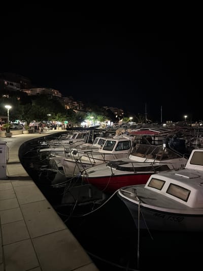 The image captures a nighttime scene at a harbor in Općina Brela, Croatia, featuring a row of moored boats along a paved walkway. The boats, varying in size and design, are illuminated by artificial lights from the shore and street lamps lining the promenade. Several boats are white with red accents, while others are primarily white with dark hulls. The water is dark and reflects the lights. In the background, buildings are visible on a hillside, with lights indicating occupancy. Trees and some foliage are present along the walkway, interspersed with streetlights. A few people are seen walking on the promenade, and some are gathered at tables in an outdoor seating area of a restaurant or cafe. The overall atmosphere is tranquil and picturesque, typical of a coastal town at night.