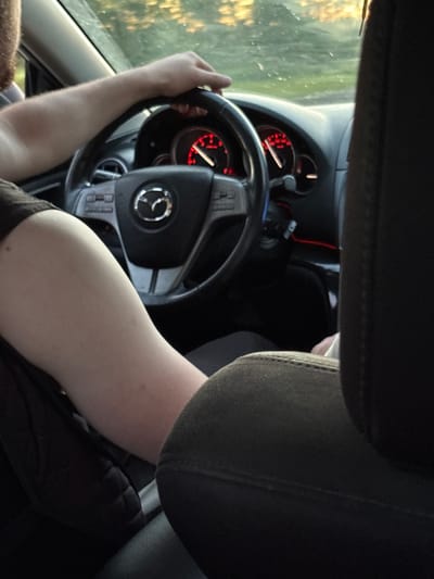 The image shows the interior of a car, with a person's arm and hand on the steering wheel. The steering wheel has a Mazda logo in the center and red illuminated gauges behind it. The dashboard is visible, along with part of the car's interior. The image is taken from the passenger's seat, looking towards the driver. The lighting suggests it is daytime, possibly late afternoon or early evening, with sunlight filtering through the windshield and reflecting on the dashboard. The background visible through the windshield is out of focus but appears to be a natural outdoor environment with trees. There is no indication of the specific location within Вяйнянскі сельскі Савет, Belarus, based on the visual information provided.