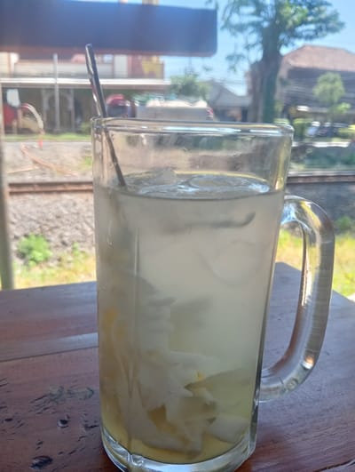 A glass mug filled with a clear beverage and ice sits on a wooden table. The mug has a handle and a straw is visible within the drink. In the background, there are train tracks and buildings, suggesting an outdoor setting. The scene appears to be in Sruni, Indonesia, based on the context provided. The lighting suggests it is daytime.