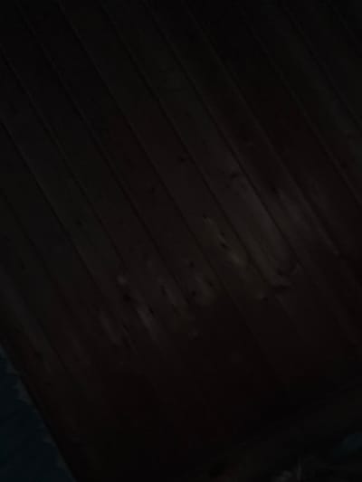 The image is a close-up, dimly lit shot of wooden planks, likely a wall or ceiling, with a dark, grainy texture. The planks are oriented diagonally from the top left to the bottom right of the frame. There are faint lighter markings on some of the planks, possibly due to wear or damage. A small portion of a dark, textured surface, possibly fabric or upholstery, is visible in the lower left corner, extending from the bottom edge of the frame. The overall impression is one of a poorly lit, possibly abandoned or neglected interior space. No specific details suggest a location within Shebarshino, Russia, beyond the provided context.