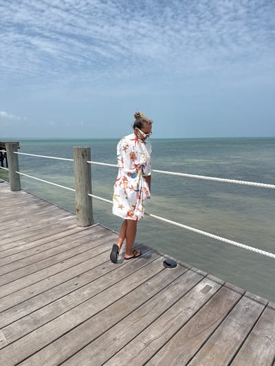 The scene depicts a single female subject standing on a wooden pier extending over ocean water in Zanzibar, Tanzania, under a bright, partly cloudy sky. The subject is oriented with her body slightly angled towards the left of the frame and her head bowed, looking down and to her right. She wears a white, short-sleeved dress or cover-up patterned with various colorful sea creatures including starfish and shells. Her hair is pulled back into a bun, secured with a yellow tie. White, angular-framed sunglasses with a thick white chain are worn on her face. Her footwear consists of black flip-flops, the right one featuring a gold-colored emblem. The pier is constructed from aged, light brown wooden planks and features a white rope railing supported by round wooden posts along its edge. The water below the pier is calm, appearing light greenish-brown, suggesting shallowness or suspended sediment. In the far left background, additional wooden structures of the pier are visible. A small, dark, dome-shaped object is located on the pier deck to the right of the subject's feet. The sky above is light blue with scattered white cumulus clouds.