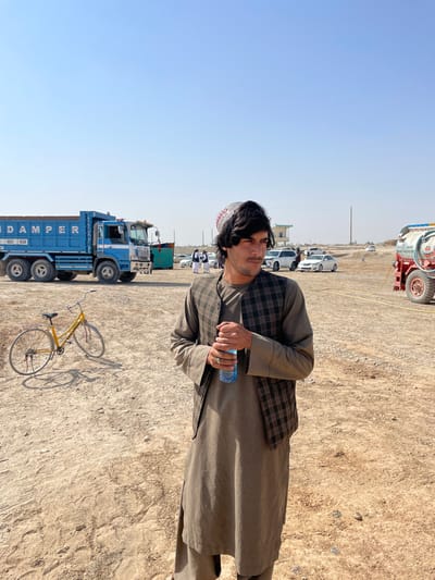 The image depicts an open, unpaved area in the city of Pir Dust, Afghanistan, under a clear, bright blue sky.
In the foreground and slightly right of center, a young adult male stands, oriented slightly rightward with his gaze directed to the right. He has dark, wavy hair, a thin mustache, and wears a light-colored traditional cap with red and white patterns. His attire consists of a long-sleeved light brown or tan shalwar kameez and a dark brown and tan plaid vest. He holds a clear plastic water bottle with a blue cap in both hands near his chest. A dark string or necklace is visible around his neck, and he wears a ring on his left ring finger.
To the left of the main subject, a yellow bicycle with fenders and a rear rack is parked on its kickstand on the dusty ground. In the mid-ground, a large, light blue dump truck with "DAMPER" visible in white lettering on its side is parked. Behind the dump truck, several individuals in white traditional attire are visible. Further in the mid-ground and background, multiple white sedans and SUVs are parked. To the far right, a red truck with a large white cylindrical tank, marked with numbers, is partially visible, with a yellow hose extending from it. In the distant background, a small light-colored building with a green roof and several vertical poles are present. The ground is dry, light brown earth with visible tire tracks and sparse debris, consistent with an unpaved outdoor area or temporary lot. The scene is illuminated by bright daylight.