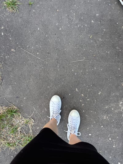 A high-angle shot captures a person's feet, clad in white low-top canvas sneakers, standing on a gray asphalt surface. The shoes have white laces tied in bows. A black skirt or dress is visible on the right side of the frame, partially obscuring the right leg. The asphalt is speckled with small white and gray particles and features scattered dry grass blades and small tufts of green grass on the edges of the frame. The overall scene suggests an outdoor setting, possibly a street or a park path, during the daytime given the lighting. There is no visible text.