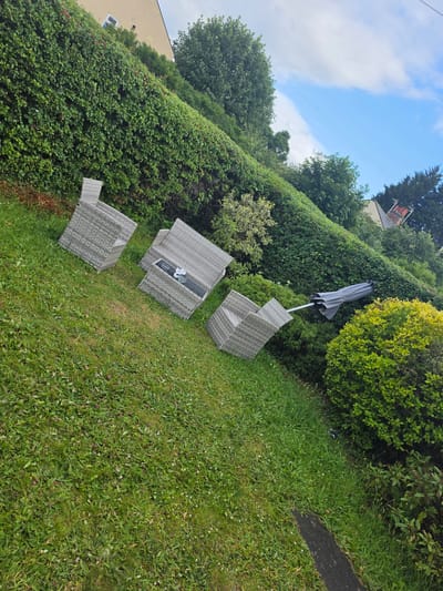This image depicts an outdoor residential garden in Shotton, United Kingdom, captured during daylight under a sky with scattered white clouds. The scene is characterized by a grassy lawn sloping upwards towards a tall, dense green hedge that forms a boundary. Arranged on the grass are three pieces of grey wicker outdoor furniture: two single-seater chairs and one combined bench-table unit, the latter holding a small white decorative object. A collapsed grey patio umbrella is lying horizontally on the grass, separated from its base, near the furniture. Various green bushes and trees are present throughout the garden, including a bright green, possibly variegated, shrub in the foreground. In the background, above the hedge, a light-colored residential building with a visible window and a section of its roof is present. A single utility line traverses the upper portion of the sky, and a distant building's chimney is partially visible against the sky in the upper right. The lawn contains visible grass clippings and assorted weeds. No individuals are present in the image.