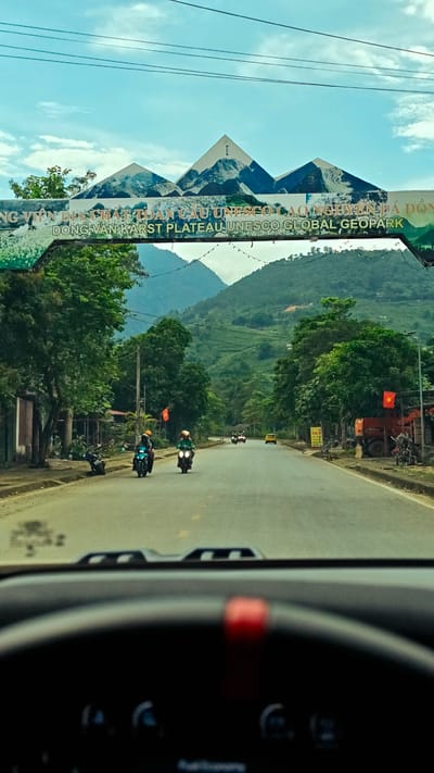 The photograph, taken from inside a vehicle, captures the view through the windshield of a straight road entering the Minh Tân Commune, Vietnam, gateway of the DONG VAN KARST PLATEAU UNESCO GLOBAL GEOPARK. The road is lined with trees and local buildings. Two motorcycles are proceeding down the road towards the camera with others further down the road, along with one taxi. The sky is blue and there are mountains in the distance. The dashboard of the vehicle is visible.