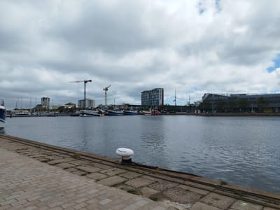 The image depicts a harbor scene in Cherbourg-en-Cotentin, France, on a cloudy day. In the foreground, a paved walkway made of rectangular stones runs along the edge of the water. A large white bollard with black trim, labeled "CHERBOURG", is situated on the walkway. Thick cables are laid on the stone surface. The water of the harbor is calm with small ripples.
Across the water, a line of docked boats is visible. These appear to be a mix of sailboats and motorboats, with masts of the sailboats extending upwards. Beyond the boats, a cityscape unfolds, characterized by several multi-story buildings. Prominent among these are two construction cranes, one blue and red and another yellow, indicating ongoing construction. One building prominently displays the name "Mercure Hotel". Another structure in the background has the signage "Les Eléis".
The sky is heavily overcast with large, grey clouds, though patches of lighter sky and possibly some blue are visible. A single bird is captured flying in the upper left quadrant of the sky. The overall scene suggests a functional port area with both maritime activity and urban development.