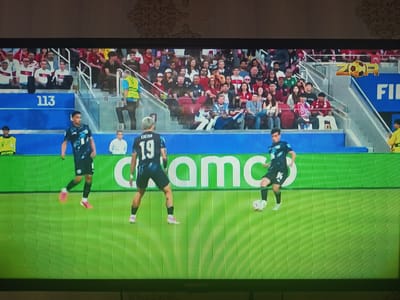 This is a photograph of a television screen displaying a soccer match. Three male soccer players in dark jerseys and shorts are on a vibrant green field. One player in the foreground is wearing jersey number 19. The other two players are further back on the field. In the background, spectators are seated in tiered stadium seating. The stadium is decorated with blue and white banners and signage, including the number "113" and a logo featuring a soccer ball. There is also a large white advertisement with the letters "amc" prominently displayed behind the players. The image is captured from a slightly elevated angle, and the lighting suggests an indoor or evening game. The scene is presented as a digital representation of a live event, likely from a video game or broadcast. There is no visible indication of Hakkulabad, Uzbekistan.