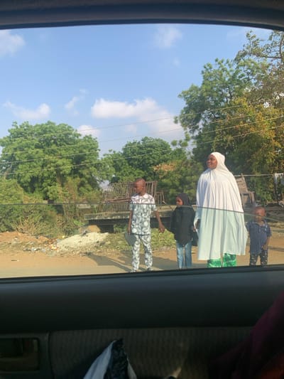 The image depicts a roadside scene in Abobo, Nigeria, viewed from the interior of a vehicle. Four individuals are present: one adult female and three children.
The adult female, positioned to the right of the group, wears a white head covering and a long white garment over a green patterned skirt. She looks towards the left. To her left stands a boy, approximately 8-10 years old, dressed in a white two-piece outfit with a black geometric pattern. He holds a dark, rectangular object in his left hand and appears to hold the hand of a smaller girl to his right. This girl, approximately 5-7 years old, wears a black head covering and black top over blue jeans, looking forward. To the adult female's immediate right is a younger boy, approximately 3-5 years old, wearing a dark blue polo shirt and patterned trousers, looking downwards.
The ground beneath the individuals is a dirt and gravel mix. To the left, refuse and debris are scattered on the ground. In the mid-ground, behind the people, are dense green trees and bushes, with utility lines horizontally traversing the scene. A concrete or stone structure, possibly a culvert or bridge, is partially visible, behind which metal frames are stacked. The sky above is clear blue with sparse white clouds, illuminated by bright daylight. The foreground shows the dark interior trim and window frame of the vehicle from which the photograph was taken.