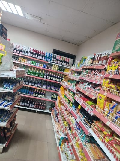 Aisles of a grocery store are shown. Shelves are stocked with various items, including bottled drinks, snacks, and packaged goods. The shelves have red price tag holders. There is a small window that shows some light from the outside. The aisles are white and the floor is tiled.