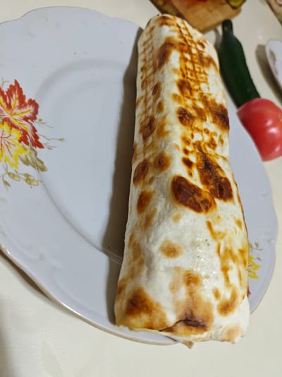 A close-up, overhead shot shows a grilled wrap on a white plate with a floral design. The wrap is golden brown and appears to be made of flatbread, with a checkered grill pattern visible on its surface. To the right of the wrap, a red tomato and a dark green cucumber are visible. The setting appears to be a kitchen or dining area.