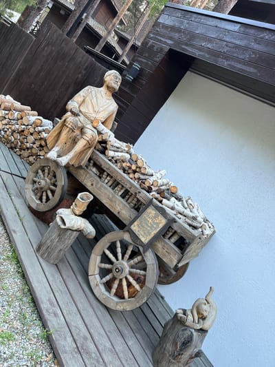 A wooden sculpture of a man sitting on a wooden cart filled with firewood is displayed outdoors. The cart has two large spoked wheels. To the left of the cart, there is a large pile of cut logs. In front of the cart, there are wooden supports for the cart's axle, and a smaller wooden sculpture of a cat resting on a log. The scene appears to be in Tal'tsy, Russia, as suggested by the architectural style of the building in the background, which features wooden siding and a dark-pitched roof. The ground is covered with a wooden walkway and gravel. The lighting indicates it is daytime.