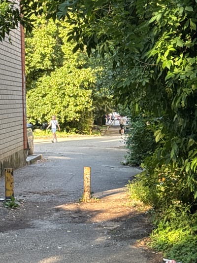 The image depicts a sunlit outdoor scene with a paved pathway bordered by lush green foliage on the right and a brick building on the left. In the background, a group of people are walking along the pathway, and a young person in a light-colored shirt and shorts is walking towards the foreground. Two yellow bollards are positioned along the pathway. The scene appears to be located in a residential or park area within the city of Verkhnerusskoye, Russia.