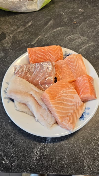 A top-down view shows a white, round plate filled with various raw fish fillets, resting on a dark, mottled countertop or table. The plate features a subtle blue floral pattern visible on its rim in several areas.
The majority of the plate is occupied by bright orange-pink salmon fillets, characterized by their distinct parallel fat lines. There are approximately five pieces of salmon, varying in size and shape, suggesting they have been cut from larger fillets. Alongside the salmon, there are at least two pieces of white fish fillets, which are paler in color and have a smoother, less textured appearance compared to the salmon. These white fish pieces are flatter and appear to be from a different type of fish, such as cod or similar.
The dark surface beneath the plate exhibits a textured, somewhat streaky pattern, resembling dark stone or a laminate material. On the far left of the image, a green and white partially visible bag or packaging can be seen, with some faint, indistinct text printed on it which is unreadable.
The arrangement suggests the fish are prepared and ready for cooking, possibly for a meal in an indoor kitchen setting, in Philippeville, Belgium. No other specific location or time details are discernible from the image.