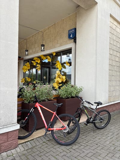 The image depicts the facade of a building in Novinki, Istrinsky District, Russia, specifically at "Pavlovo Podvorye, Building 4." Two bicycles are parked on a paved walkway in front of the building's entrance. The building has a beige brick facade and a white pillar. Hanging lanterns are visible under an overhang above the entrance. Large, artificial yellow flowers are decorating the windows. In front of the windows are large planters containing green and variegated foliage. One bicycle is a bright coral color with black wheels, and the other is a dark gray with green accents and black wheels. Both bicycles are standing upright, supported by their kickstands. The walkway is made of grey interlocking pavers.
