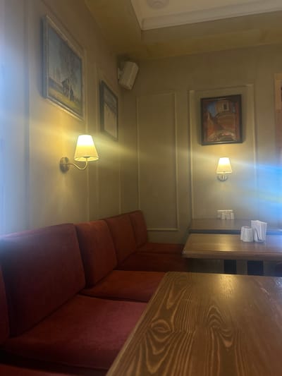 The image depicts the interior of a dimly lit dining establishment, likely a cafe or restaurant, in the city of Vyatskoye, Russia. In the foreground, a dark wooden table with a prominent wood grain pattern occupies the lower portion of the frame. Behind it, a lighter wooden table is visible, set with a stack of white napkins, salt and pepper shakers, and two small patterned cups.

A long, deep red upholstered banquette seating area stretches along the left and rear walls of the room, creating a cozy, intimate setting. Mounted on the wall above the banquette are two wall sconces with cream-colored lampshades, casting a warm, ambient glow.

The walls are painted a neutral, muted tone, and several framed paintings adorn them. On the left, a landscape painting featuring a rustic wooden house and a tree is hung. To its right, a smaller, darker framed picture is visible. Further to the right, above the second table, another painting showcases a building with a dome, rendered in earthy tones.

A white speaker is mounted on the wall in the upper left quadrant of the image. The ceiling appears to be a pale yellow color with subtle molding details. The overall atmosphere suggests a quiet and possibly historic dining space, characteristic of establishments found in older Russian towns like Vyatskoye.