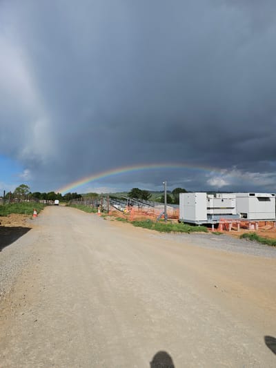 The image depicts a rural construction site near Lacock, United Kingdom. A dirt road runs along the right side, with a fence and some orange construction barriers lining the edge. Large, white, prefabricated units are visible adjacent to the road. Across from the road, there's a full rainbow arching across a partly cloudy sky, with dark storm clouds dominating the upper left and brighter skies near the lower left. The rainbow's endpoint seems to be in the vicinity of a row of trees and construction area in the distance. Shadows suggest that the image was taken during the day. No people are visible in the image.
