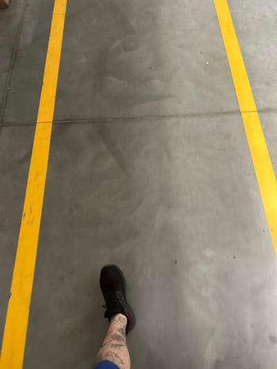 The image shows a person's legs and feet from a first-person perspective, standing on a gray concrete floor. Two parallel yellow lines are painted on the floor, running vertically from the bottom of the frame towards the top. The person is wearing a black athletic shoe with red accents and has tattoos visible on their lower left leg. The environment appears to be an indoor space, possibly a warehouse or industrial area, given the concrete flooring and painted lines. There are no other people or objects clearly visible in the immediate vicinity. The location in Białki, Poland, is not visually indicated by any landmarks or signage within the image.