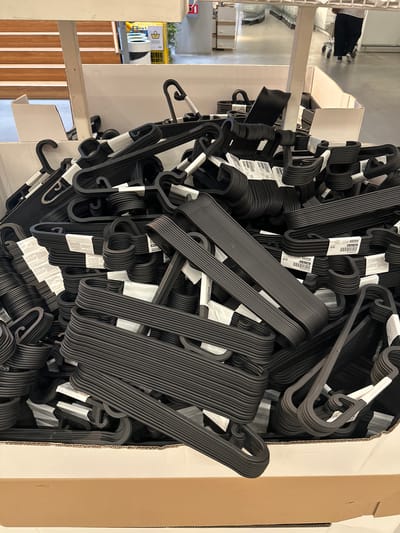 A close-up, overhead shot captures a large, overflowing cardboard box filled with black plastic clothes hangers. The hangers are stacked haphazardly, with some neatly aligned and others jumbled together, creating a dense, textured surface. Many of the hangers have white paper tags attached, some of which are partially obscured by the surrounding hangers. One visible tag reads "Article Number 104.184.86". The lighting suggests an indoor setting, possibly a warehouse or retail store, with diffuse light illuminating the scene. The composition focuses entirely on the mass of hangers, with the edges of the cardboard box framing the bottom and right sides of the image. The overall impression is one of abundance and bulk packaging. The setting appears to be a retail environment, given the packaging and the nature of the items. There are no people visible. The image is captured from a high angle, looking down into the box. The background, though out of focus, shows a continuation of what appears to be shelving or more product in a commercial space. The color palette is dominated by black, white, and the brown of the cardboard.