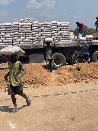 A truck is heavily laden with stacked sacks, likely cement. Two individuals are atop the truck, seemingly unloading the sacks. Another person on the ground carries a sack on their shoulder. Dust covers the people and the sacks. The environment appears to be a dusty road with a clear sky in the background.