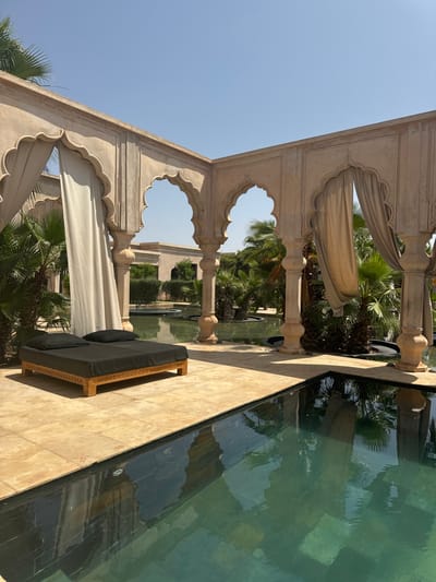 The image shows an outdoor patio in Ouled Jelal, Morocco, at coordinates 31.664 latitude, -7.87 longitude. The patio features a rectangular swimming pool with a light green, clear water surface. A wooden lounge chair with black cushions is placed to the left of the pool. The patio is enclosed by a series of archways formed by stone columns, draped with light brown fabric. There are palm trees and other lush greenery visible outside the patio. The sky is clear and blue.