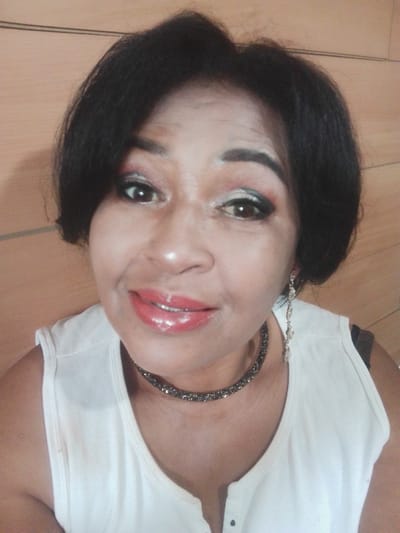 A medium shot shows a Black woman posing for a selfie in front of a wood-paneled wall. The woman has short, dark hair and is wearing bright red lipstick, eye makeup, and a white tank top. She is also wearing a sparkling choker necklace and a dangling earring in her right ear. The image appears to be taken indoors.