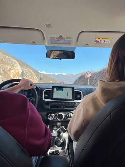The scene depicts the interior of a vehicle traveling on a road in Miločani, Montenegro. The perspective is from a rear seat, facing forward through the windshield on a clear, sunny day with a bright blue sky.

Two individuals occupy the front seats. The driver, positioned on the left, is wearing a dark red long-sleeved garment. Their right hand, adorned with a red string bracelet, is on the steering wheel. The front passenger, on the right, is partially visible from behind, exhibiting long brown hair and wearing a light brown hoodie.

The vehicle's interior features a black dashboard. A central infotainment screen displays a navigation application, showing a route with text in Cyrillic, indicating a destination approximately 11 minutes away. Below the screen are climate controls with two circular knobs and buttons. A manual gear shifter with a black knob is located on the center console, which also contains two USB ports, one with a white cable plugged in, and a snack wrapper. The passenger-side sun visor is lowered, revealing an "AIRBAG" warning label.

Outside the vehicle, a paved road extends ahead through a mountainous valley. The adjacent mountainsides are partially covered in snow and ice. Distant mountain peaks are entirely snow-capped. Road signage visible includes a circular speed limit sign on the right, indicating "50" (km/h), and a triangular warning sign on the left. A red car is observed in the distance on the road, heading in the same direction.