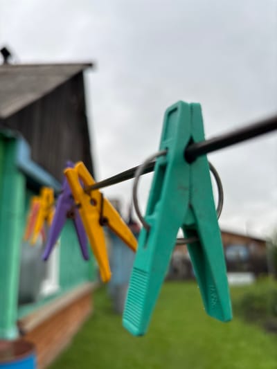 A close-up, shallow depth of field shot focuses on a green plastic clothespin clipped to a clothesline, which is stretched horizontally across the frame. The clothespin is in the foreground and is in sharp focus, showing a ribbed texture on its lower arms. Behind it, slightly out of focus, are several other clothespins of different colors—purple and yellow—also attached to the clothesline.

In the background, a weathered wooden house with a dark roof is visible on the left, partially obscured by the clothespins. The house has a green painted section on its lower visible part. To the right and further in the background, a blurred green lawn and some out-of-focus structures or vegetation are present. The sky above is overcast and light gray. The overall impression is of an outdoor setting, likely a backyard or laundry area. The image does not provide any explicit information to indicate it is specifically located in the city of Gonja, Russia.
