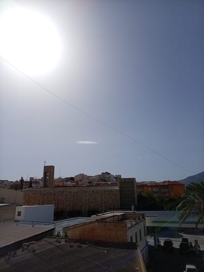 This media file captures a daytime scene in Olula del Río, Spain, under a bright, sunny sky. The sun is prominently visible in the upper left quadrant, suggesting it is mid-morning or early afternoon. The weather appears clear and warm, with a faint, wispy cloud visible in the sky.
The image focuses on a town or city landscape. In the mid-ground, a prominent stone church tower with a cross on top stands tall, flanked by other buildings with red-tiled roofs, characteristic of Spanish architecture. Below the church, a large stone-faced building is visible, possibly part of the church complex or another public building. Further to the right, an orange apartment building and other residential structures are nestled amongst trees.
In the foreground, several rooftops are visible, indicating an elevated viewpoint. The roofs are made of corrugated metal and other materials. To the far right, a gas station with a canopy and cars can be seen, with a palm tree adding a tropical touch to the scene. The overall impression is that of a typical Mediterranean town on a clear, sunny day. There is no visible text in the image.