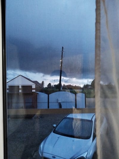 This is a medium-shot, eye-level photograph taken from indoors looking out through a window. The scene depicts a rural or suburban outdoor setting under a stormy sky. In the foreground, the bottom right corner shows the corner of a white car, possibly a hatchback, with its headlights visible. The car is parked on a gravel surface.

The midground features a corrugated metal fence, which is light blue and appears to be in two sections. Behind the fence, there are several buildings, including a white-sided house with a dark roof and a garage-like structure with large, dark doors. Further back, there are more houses with reddish-brown roofs and trees, some of which are swaying as if in strong wind. A wooden building is partially visible to the left of the garage.

The background is dominated by a dark, dramatic sky filled with heavy, ominous clouds, suggesting impending rain or a storm. A single utility pole with wires stretches across the sky, and a faint light can be seen on it. The overall mood is somber and potentially tense due to the stormy weather. The time of day appears to be late afternoon or early evening, given the dim lighting and the dramatic sky. Reflections on the window are visible, particularly a vertical, light-colored curtain on the right side of the image. No text is visible in the image. The location context is Sudzhenka, Russia.