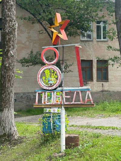 The image depicts an outdoor scene in Teploozyorsk, Russia, featuring a colorful, handcrafted sculpture. The sculpture appears to be made from repurposed materials, including tires and metal framing, and is painted in vibrant red, yellow, blue, and white. It is positioned in front of a weathered, two-story building with multiple windows. Lush green grass and trees surround the sculpture, suggesting a natural or park-like setting.
The sculpture itself is abstract, with several circular elements that resemble painted tires and a more intricate, geometric structure at the top. Some parts of the sculpture have visible text painted in white letters that read "100 ЛЕТ" which translates to "100 YEARS". The overall impression is one of folk art or a community art project. The lighting suggests it is daytime, likely during pleasant weather, as evidenced by the bright foliage and the absence of harsh shadows. There are no people visible in the image. A rusty barrel is present to the right of the sculpture, further contributing to the sense of an outdoor, possibly semi-abandoned or repurposed area.