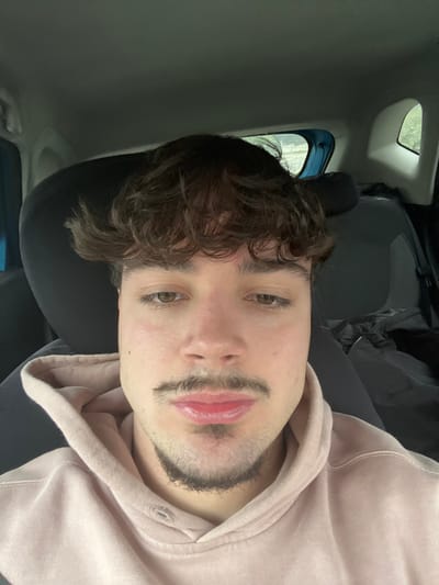 The image displays a young adult male positioned in the interior of a vehicle, facing directly forward. The subject has dark, curly hair with a prominent fringe, brown eyes, a thin mustache, and a small goatee. He is wearing a pale pink hooded sweatshirt. A dark headrest is visible behind him. To the subject's right, a dark car seat and various dark fabric items are present. A partially visible window in the upper right background reveals blurred outdoor greenery. No discernible external landmarks are visible to confirm the location as Lamego, Portugal. The lighting is diffused, consistent with an interior setting lit by ambient daylight. The subject appears to be taking a self-portrait.