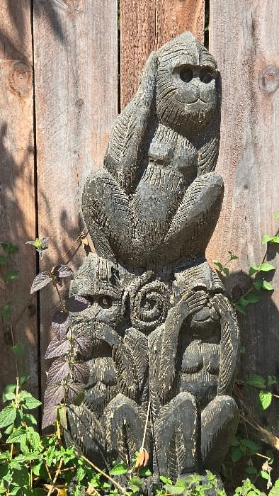 A carved, gray wooden sculpture rests on weathered wooden planks. The sculpture features textured surfaces, with a face-like design at one end and elaborate geometric carvings along its length. Green plants with small leaves and a few stems of purple-tinged foliage surround parts of the sculpture and grow between the wooden planks. Sunlight casts shadows across the planks, adding depth to the composition.