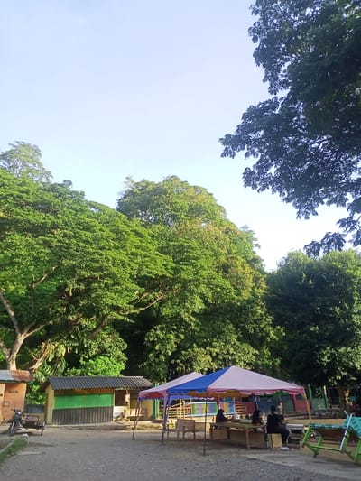 The image depicts an outdoor scene with lush green trees dominating the upper half. In the foreground, there are several small structures that appear to be shops or food stalls with colorful awnings. A blue and pink tent provides shade over a seating area with wooden tables and benches, where several people are seated. To the left, a motorcycle with a small trailer is parked near a building. The ground is covered with gravel. The sky is clear and bright. The scene suggests a relaxed outdoor gathering or a market in a tropical environment. The location is identified as Geundring, Indonesia.