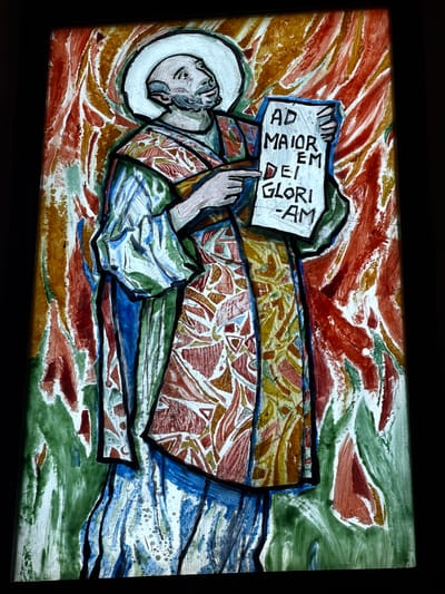 The image shows a stained glass artwork depicting a religious figure lying down, possibly deceased, with a halo around the head. The figure is holding a sign that reads "AD MAIOR DEI GLORI AM". The background consists of a mix of red, orange, green, and brown hues, with brushstroke-like textures, giving the impression of an ethereal or abstract setting. The figure is clothed in robes of various patterns and colors. The style is reminiscent of medieval or renaissance religious art, with bold outlines around the figures and design elements. The location is Grad Opatija, Croatia, this is based on common themes in religious art found in the region.
