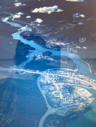 The image is an aerial view of a coastal town or city situated at the confluence of a river and the sea. The scene depicts a developed urban area with a grid of streets and buildings, some of which appear to be large, possibly commercial or industrial structures, alongside denser residential areas. Roads curve and weave through the landscape, connecting the built-up areas to the surrounding natural environment.
A prominent feature is the body of water, a river or strait, that separates the urban development from a mountainous or heavily forested landmass on the left side of the image. The water is a clear, bright blue in some areas, suggesting depth or clarity, and turns a lighter, more milky blue in other sections, perhaps indicating shallower waters, sediment, or reflections. There are also patches of white, cloud-like formations visible over the mountains, suggesting a partly cloudy sky or perhaps snow at higher elevations.
The time of day is not explicitly clear, but the lighting suggests it could be daytime, possibly with some haze or atmospheric conditions affecting the clarity of the image. The overall impression is one of a vibrant, populated area nestled within a scenic natural landscape. The image is a still photograph, and no specific activity or event is captured, nor are any people or distinct objects of interest readily identifiable beyond the general features of the landscape and urban development. There is no visible text in the image. The location context provided is Kuta Pungwa, Indonesia, which, if accurate, would place this scene within a tropical or subtropical environment.