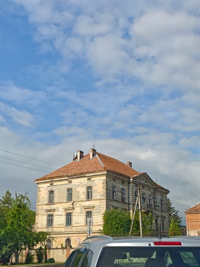 In the city of Monastyryska, Ukraine, a multi-story building with a weathered, light-colored facade and a terracotta tile roof stands under a bright blue sky dotted with white clouds. The building exhibits architectural details such as multiple windows with dark frames and a prominent triangular pediment above the central upper-floor windows. A small balcony is visible on the second floor, to the right of the building's center. Power lines crisscross the sky in front of the building. The foreground of the image shows the rear of a silver car, its roof and antenna visible. Lush green trees and foliage surround the base of the building and extend into the scene on the left. A low, cream-colored wall borders the property in front of the trees. The scene appears to be an outdoor, daytime setting.