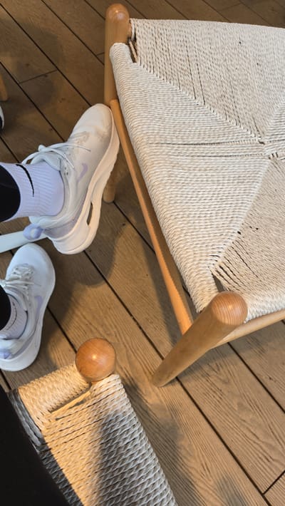 The image shows a close-up view of two white athletic shoes, one in focus and one slightly out of focus, resting on a light-colored wooden floor. A light beige woven chair with wooden legs is partially visible on the right side of the frame. The shoes appear to be in a casual setting, possibly indoors. The lighting suggests a well-lit environment, possibly with natural light. There is no indication of the specific location within Gwangmyeong, South Korea. The image primarily focuses on the footwear and the chair, creating a minimalist and clean aesthetic.