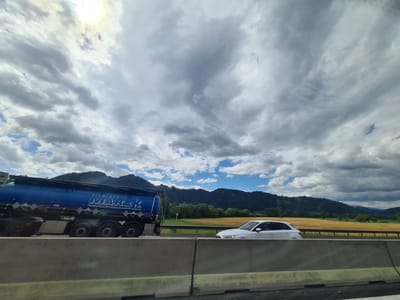 The image captures a highway scene in the vicinity of St. Stefan ob Leoben, Austria, under a dramatic sky with a mix of sun and clouds. In the foreground, a concrete barrier runs across the bottom of the frame. To the left, a blue tanker truck with "MAREK" and "FIRMENGROUP" written on its side is in motion. It has multiple axles and is likely carrying a liquid. To the right of the truck, a white sedan is also traveling on the highway. Behind the vehicles, a yellow field stretches towards a dark green, tree-covered mountainside. The sky above is filled with heavy, gray clouds interspersed with patches of blue sky and bright sunlight peeking through. The overall impression is one of travel on a clear but potentially inclement day in a rural Austrian landscape.