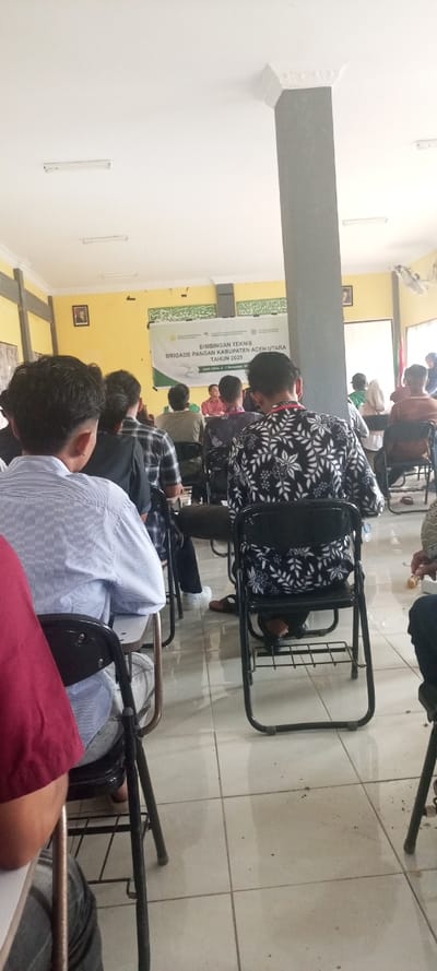 An indoor photograph depicts a seminar or training session in Matang Raya Blang Sialet, Indonesia. The vantage point is from the rear of the audience, looking towards the front of the room. Approximately fifteen to twenty individuals, predominantly men, are seated in rows on black metal folding chairs, many with attached desk surfaces, facing a presentation area.
The room features bright yellow walls, a white ceiling with multiple fluorescent light fixtures, and a light-colored tiled floor. A prominent gray rectangular pillar extends from floor to ceiling in the right midground. Decorative white molding is present along the ceiling edges. On the far yellow wall, a large white banner with green and black text is displayed. The banner reads, "BIMBINGAN TEKNIS BRIGADE PANGAN KABUPATEN ACEH UTARA TAHUN 2025," with additional text indicating "Aceh Utara, 5-7 November." Above the banner, green Arabic script is visible. A framed portrait hangs on the left wall. A speaker box is mounted on the upper left wall, and a white ceiling fan is partially visible on the upper right.
At the front of the room, several individuals are seated behind a white table, facing the audience. One person in a reddish-brown shirt is visible among them, appearing to be a speaker or instructor. Audience members are dressed in a mix of casual and semi-formal attire, including a blue and white striped shirt and a black shirt with a white floral pattern. Footwear visible on the tiled floor includes sandals and white sneakers. A small plastic bottle is visible on the floor near a chair in the midground.