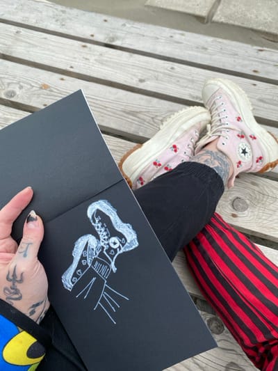The scene depicts a first-person perspective of an individual seated on a weathered wooden plank surface, likely an outdoor deck or boardwalk in Ulcinj, Montenegro. The individual's lower torso, legs, and left hand are visible. The left hand, adorned with tattoos including a trident-like symbol and a wavy line, holds open a black sketchbook. The right page of the sketchbook displays a white line drawing of two high-top sneakers, stylized with a star logo and small hearts, resembling the footwear being worn. The individual's left leg is clad in a black jogger-style pant, while the right leg wears a red and black vertically striped garment. Both feet are fitted with light pink, cherry-embroidered high-top Converse All Star sneakers featuring thick cream platforms and rugged tan outsoles. Tattoos are visible on the ankles and lower shins beneath the shoes.