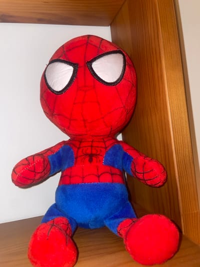 A plush toy of Spider-Man rests in a shelf. The toy is red and blue, featuring the character's classic costume design with a red head and torso, blue body, and web patterns. White eyes are prominently displayed on the head. The toy appears to be in good condition. The shelf has a white backing and a light-colored wooden frame.