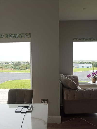 This media file shows an interior view looking out through two windows towards a rural landscape in Doirí Beaga, Ireland. The scene is set in a modern room, with a light-colored wall dominating the foreground. On the left, a large window offers a view of rolling green hills, scattered houses, and a wooden structure that resembles a playground or animal pen. The sky is overcast, suggesting a cloudy or potentially rainy day.

In the foreground, a white marble-topped table is visible with two smartphones and a charging dock placed on it. A dark brown leather chair is positioned behind the table, partially obscuring the view through the left window.

To the right, another window looks out onto a different section of the landscape, featuring a light blue building and more of the green terrain. A tufted sofa in a neutral beige fabric is positioned in front of this window, with a cushion placed on it. On a small round table next to the sofa, a potted orchid with vibrant purple flowers is displayed.

Further to the right, an electrical outlet with two plugs is mounted on the wall. A grey radiator is visible behind the sofa. The overall impression is a comfortable, modern living space with a view of the Irish countryside. There is no visible text in the image. The time of day appears to be daytime, given the natural light.
